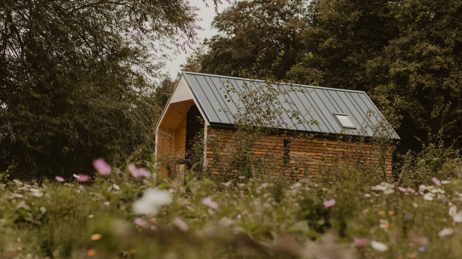 A small wooden cabin with a metal roof sits surrounded by wildflowers and trees on a cloudy day, evoking the charm of a rustic boat house.