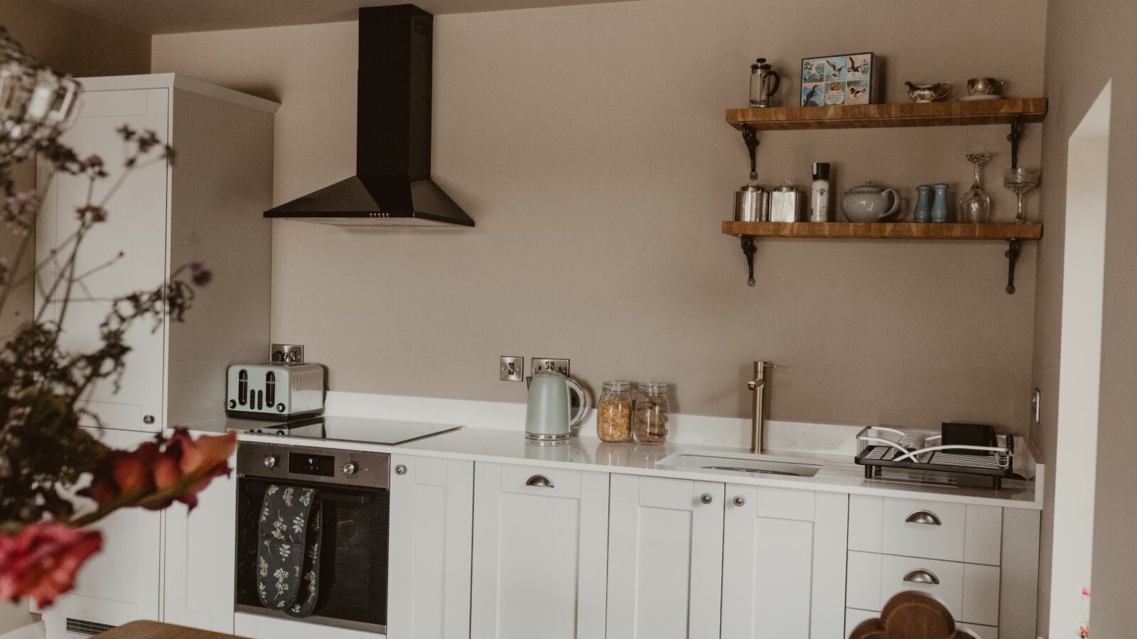 Modern kitchen in a Boat House, featuring white cabinets, a black range hood, and wooden shelves stocked with jars and bottles—perfect for prepping meals after a day of water activities. A kettle sits invitingly on the counter, ready to brew up warmth and comfort.