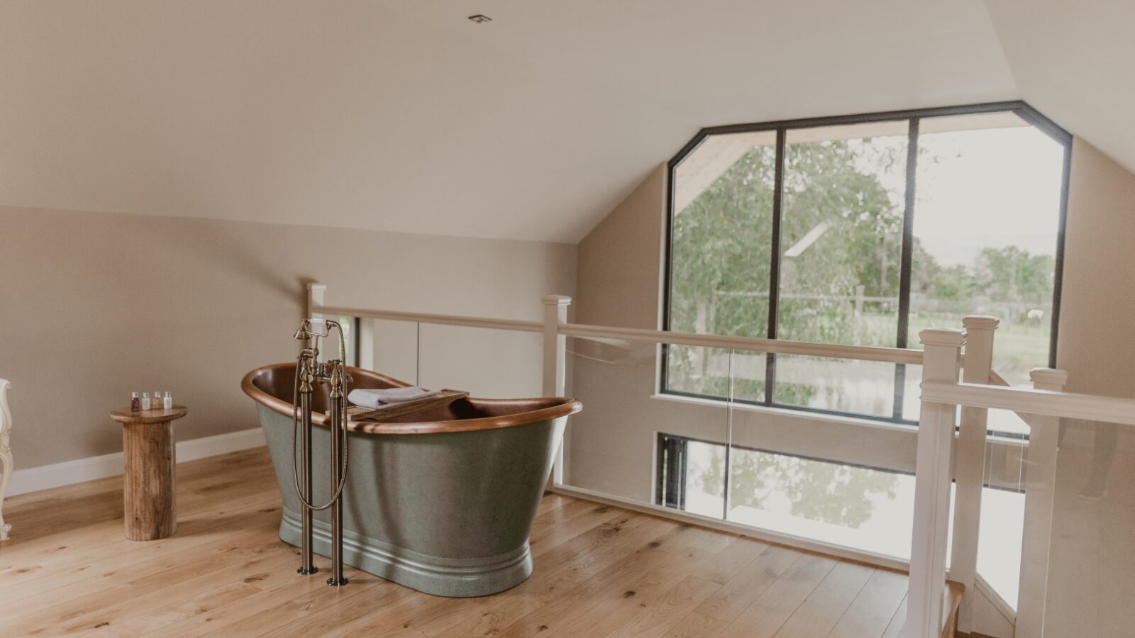 A freestanding metal bathtub on a wooden floor near a large window offers an exquisite view of trees, evoking the serene charm of a boathouse nestled by the water.