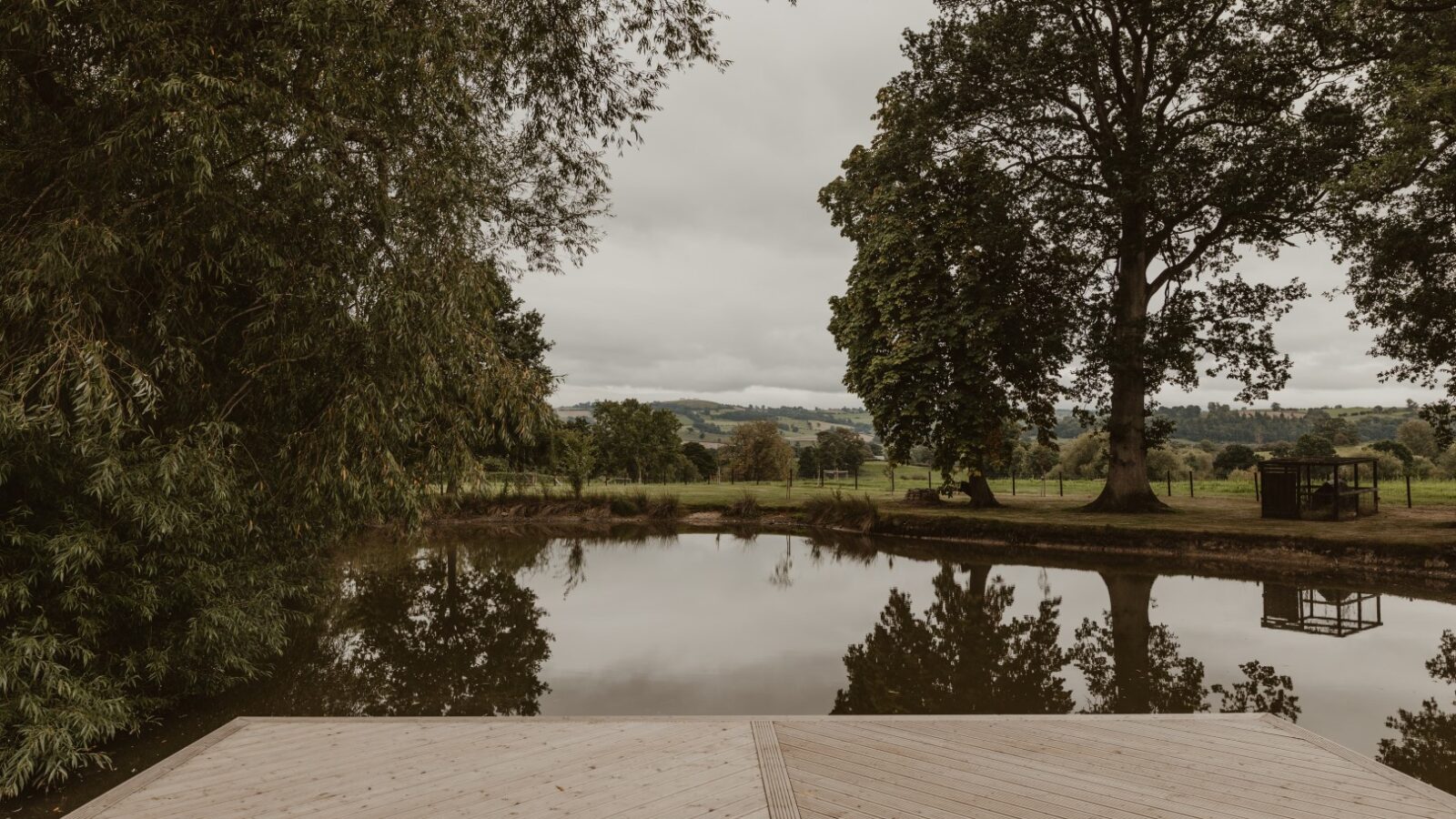 A wooden deck, reminiscent of a boat house, overlooks a serene pond surrounded by trees, with cloudy skies and a distant view of rolling hills in the background.