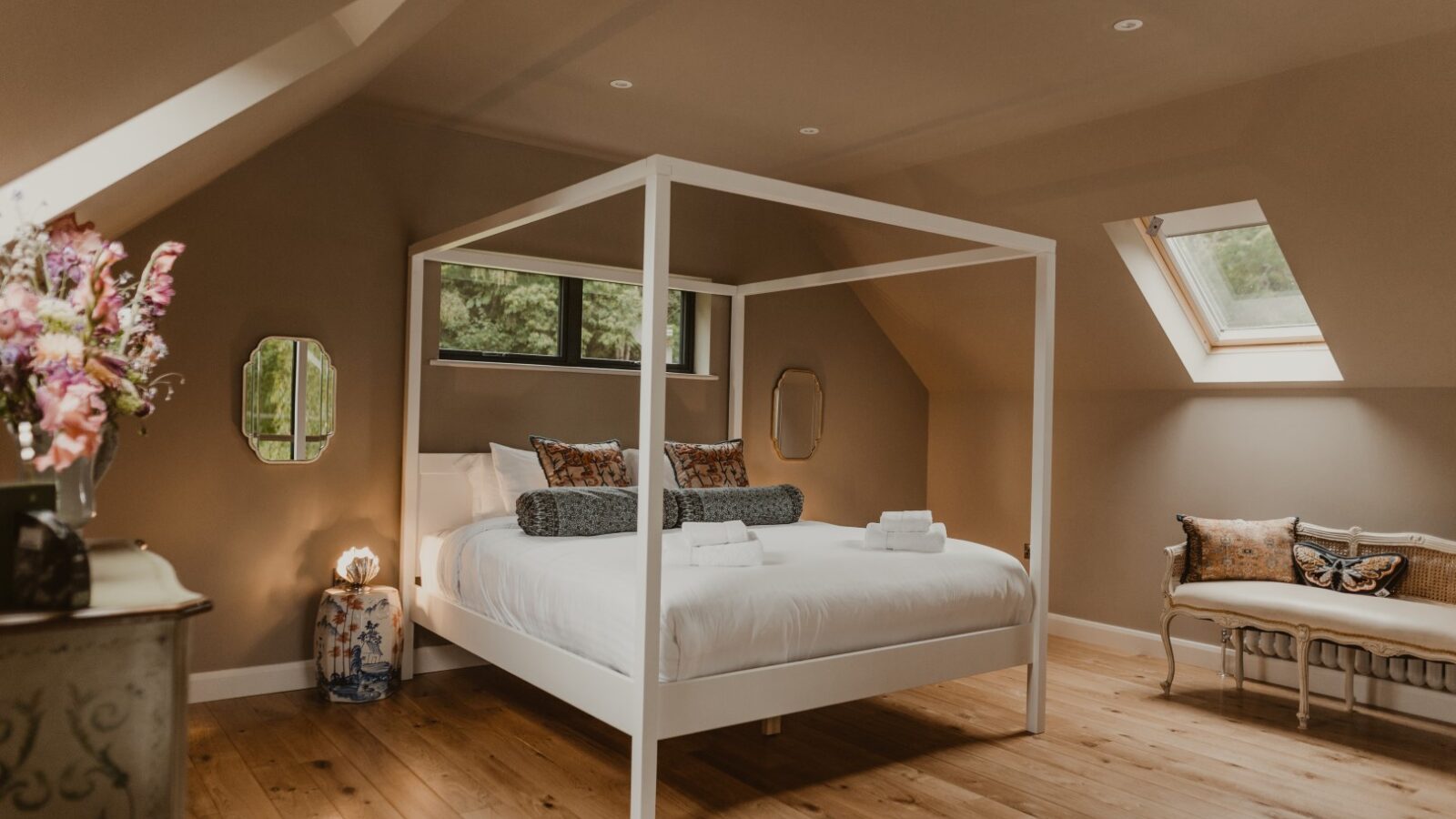 A cozy bedroom in the Boat House features a four-poster bed, skylight, decorative mirrors, flowers on a dresser, and soft lighting, perfect for unwinding after a day on the waterfront.