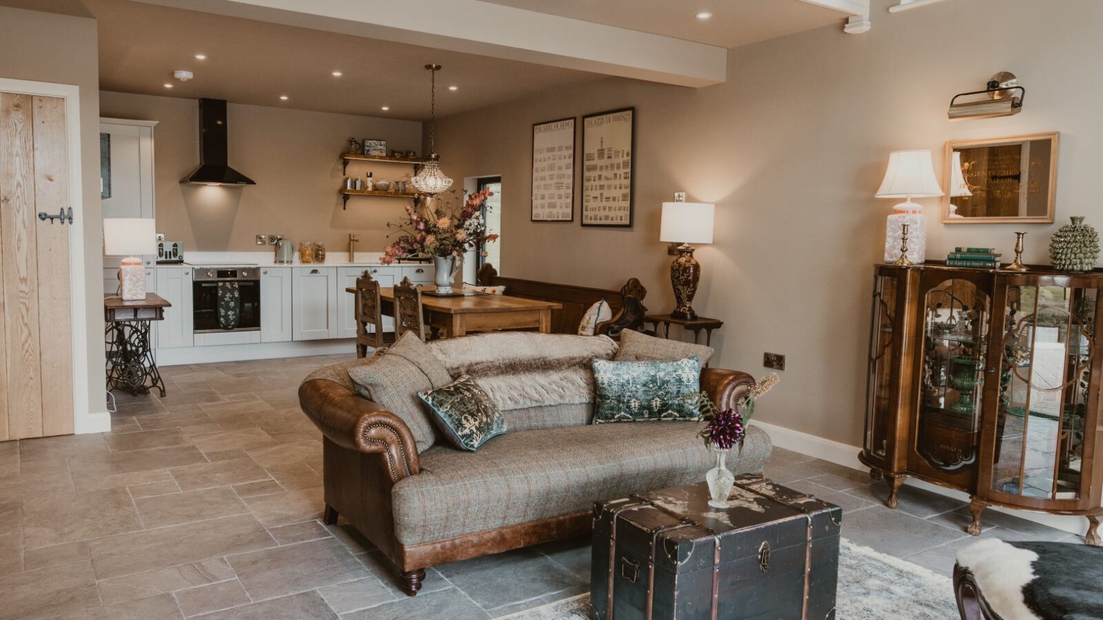 Cozy living room in The Boat House, featuring a vintage sofa, trunk coffee table, and adjacent kitchen. Decor includes lamps and framed wall art, perfect for relaxing after a day of waterfront dining or boat rentals.