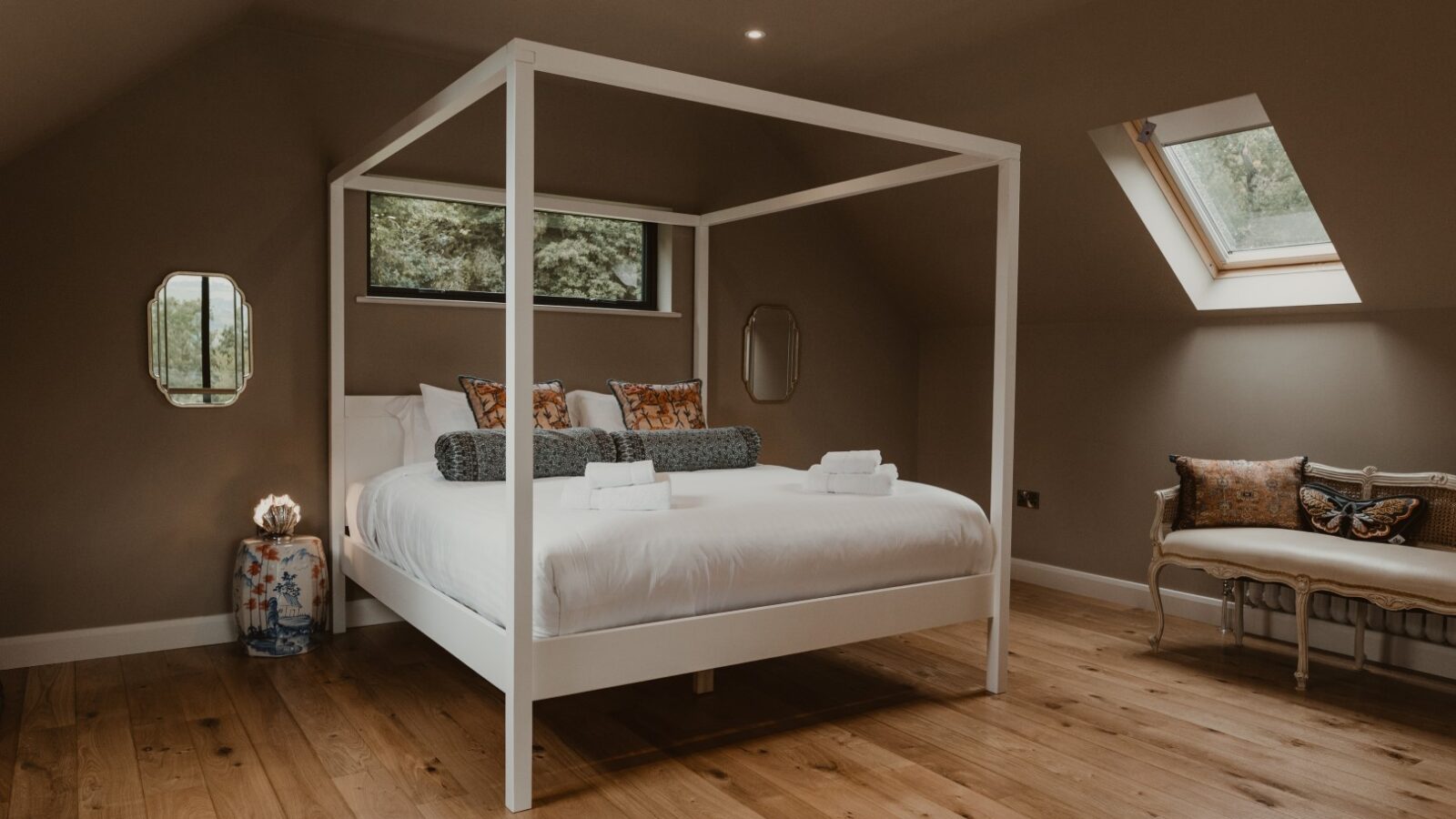 A spacious bedroom in a charming Boat House features a white four-poster bed, decorative pillows, wooden floor, skylights, and neatly folded towels on the bed.