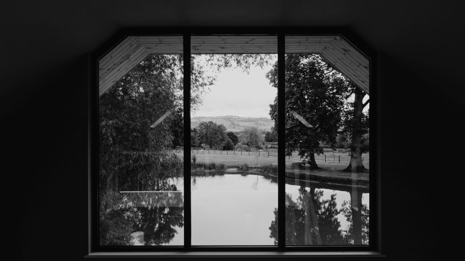 A black and white view through a window reveals a charming event venue with trees, a serene pond, and rolling hills in the distance.