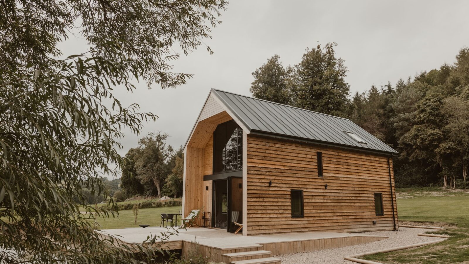 Nestled among the trees and grassy landscape, this charming wooden cabin, known as the Boat House, boasts a metal roof and features a cozy outdoor patio with chairs. It's an idyllic event venue where guests can enjoy waterfront dining in a serene natural setting.