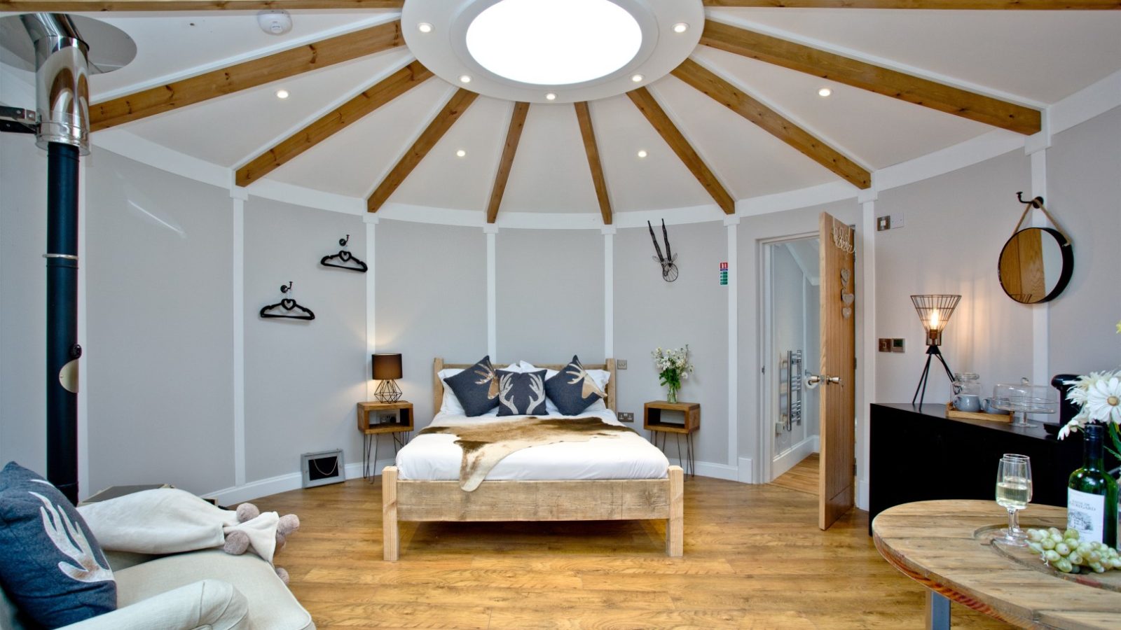 A cozy circular room at East Thorne with wooden beams radiating from a central skylight. The room features a double bed with pillows and blankets, a small round dining table, a sofa, and a black dresser. Various wall decorations, flowers, and neatly arranged accents add charm to the space.