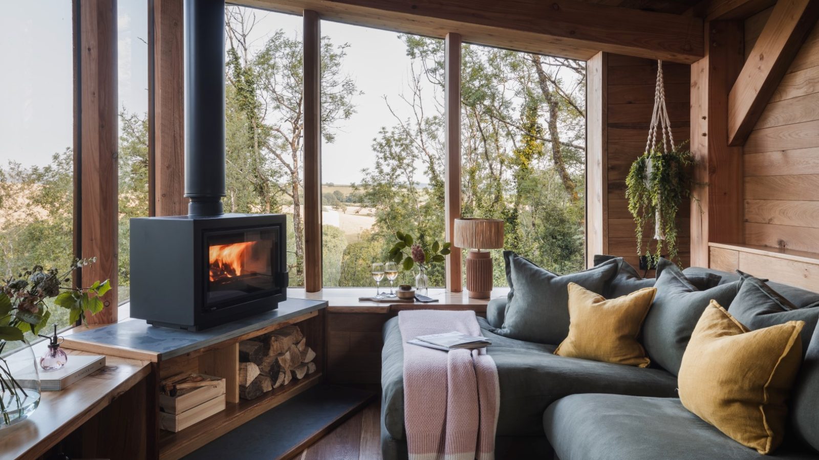 Cozy cabin interior in Wolf Wood Treehouses with a wood-burning stove, large window views, green sofa, and yellow cushions.