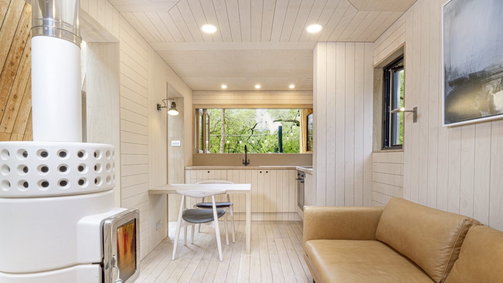 The modern minimalist living area feels like a dazzle treehouse, with light wood walls, a beige sofa, and a small dining table. Large windows immerse you in breathtaking forest views.