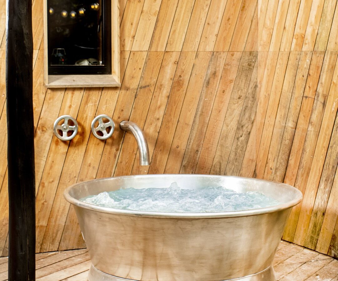 A metal bathtub brims with water in a Dazzle Treehouse-themed wooden-paneled room, illuminated by three hanging lights above and a small window on the wall.