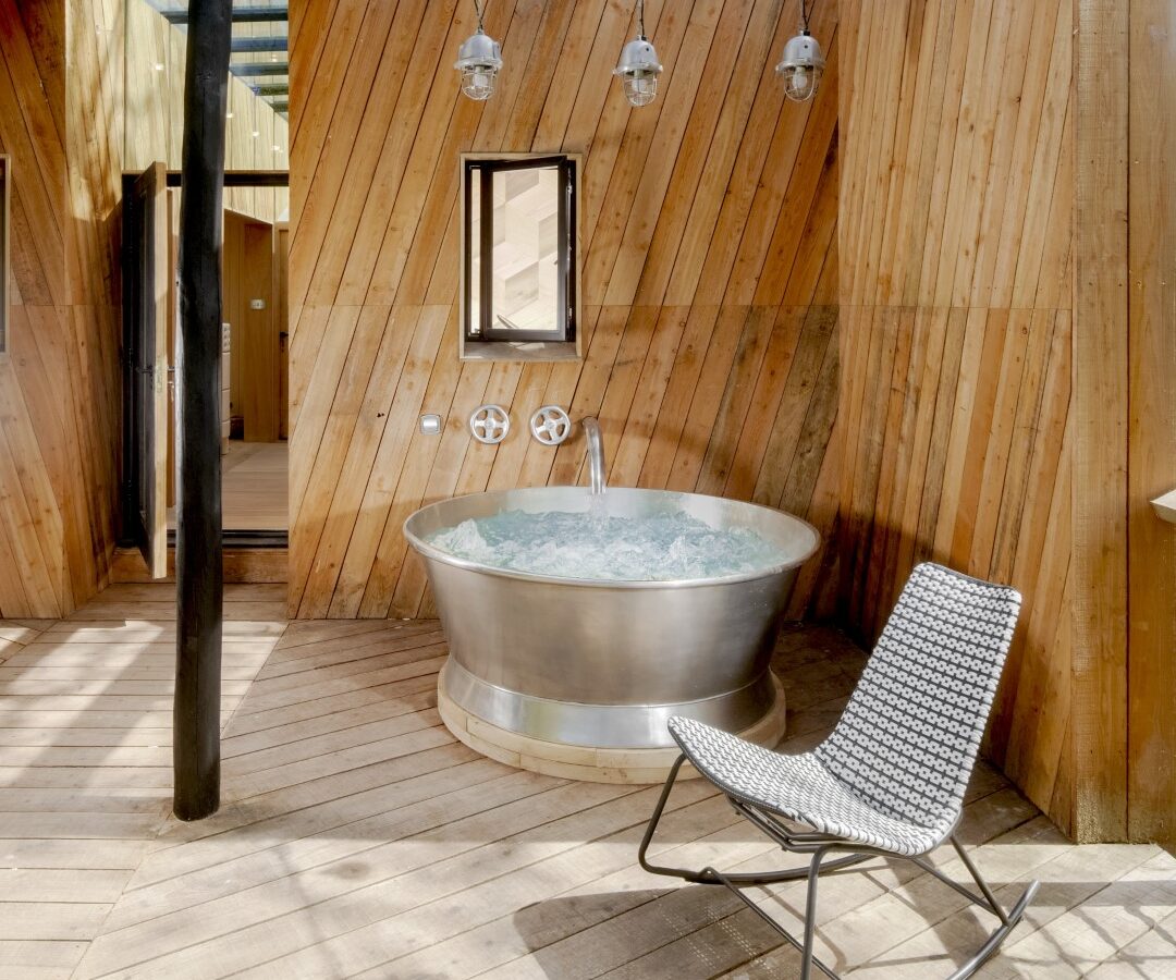 The Dazzle Treehouse features an outdoor wooden deck with a round metal hot tub, illuminated by three pendant lights. Beneath the pergola, a woven chair invites relaxation in this enchanting oasis.