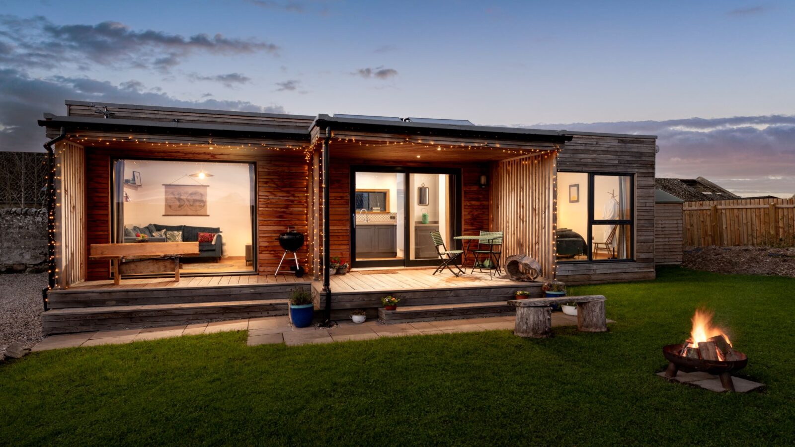 A modern, wood-clad cabin features outdoor lights, a patio with seating, a lush lawn, and a small fire pit in the foreground, reminiscent of a cozy retreat.