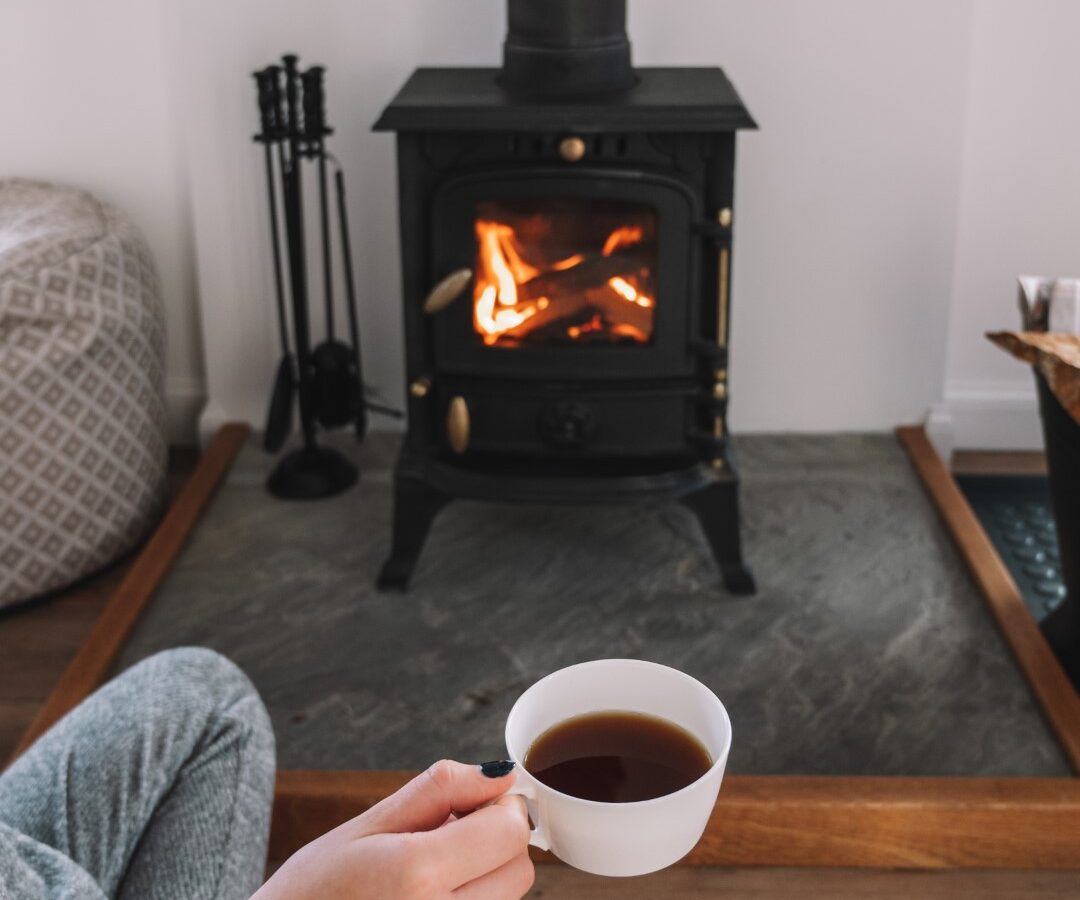 Person in gray pajamas holding a cup of tea near a lit wood-burning stove on a wooden floor, with a Frances Sketch Pad and drawing tools scattered nearby.