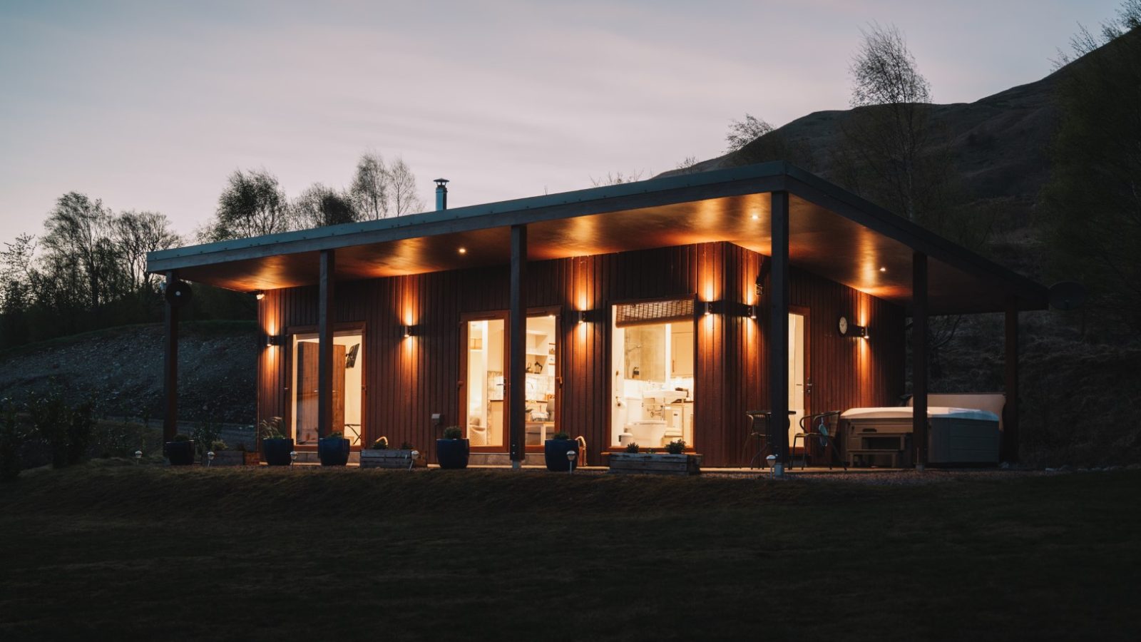 A modern, single-story house with large windows and a wooden exterior is warmly illuminated at dusk. It sits in a grassy landscape with hills in the background, reminiscent of a scene from Frances Sketch Pad. A hot tub is visible on the right side, enhancing its serene charm.