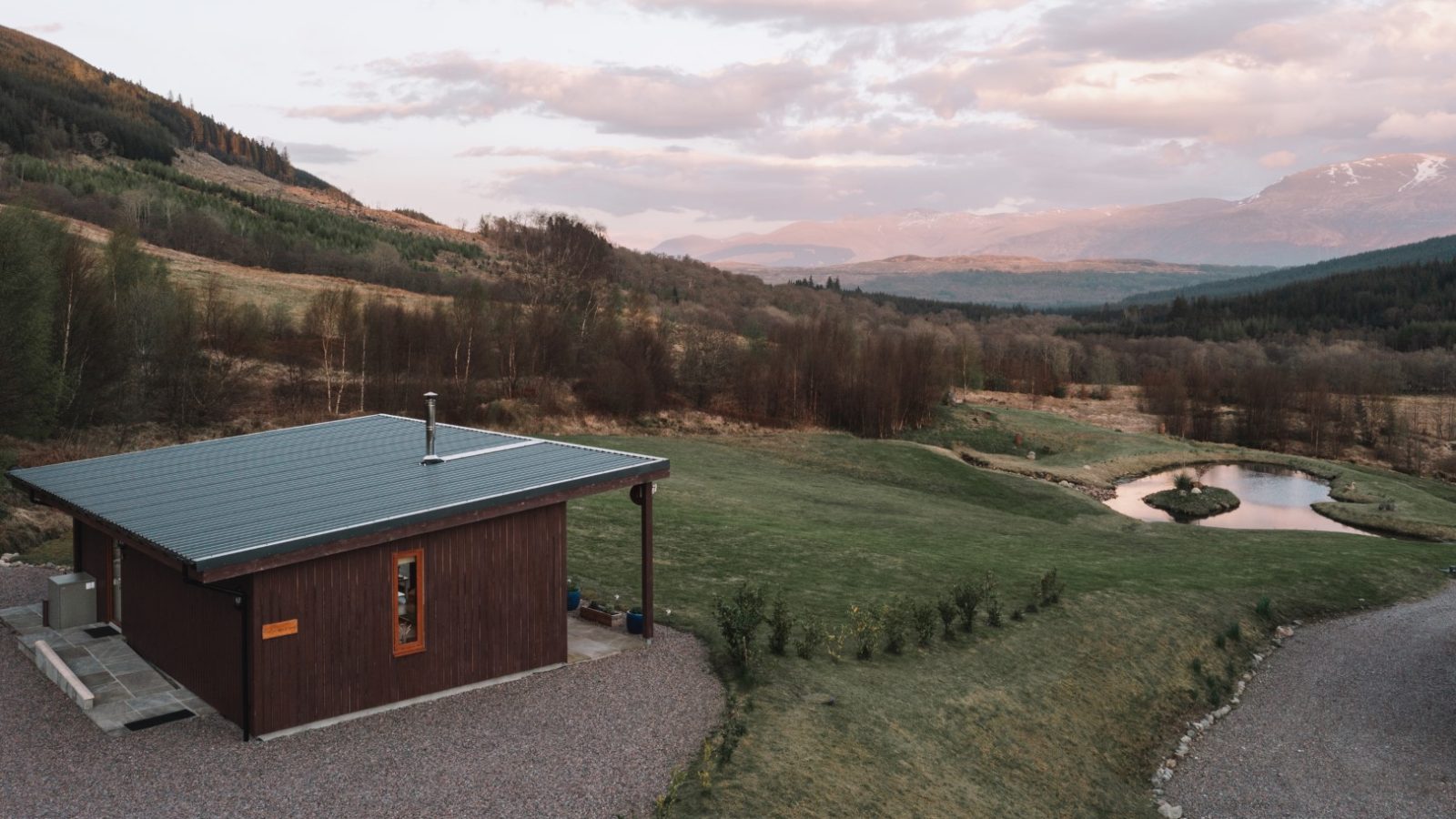 A small wooden cabin with a green roof sits in a scenic valley, as if plucked from Frances's sketch pad. Surrounding it are rolling hills, a grassy landscape, and a pond. The area is lush with trees and distant mountains under a partly cloudy sky at sunset.