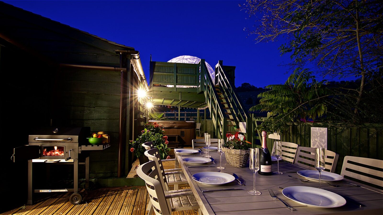 Beneath a clear night sky, Georges Getaway invites you to its outdoor dining area, featuring a table set for six and a nearby grill. Stairs lead to a cozy raised platform, perfect for savoring evening moments.