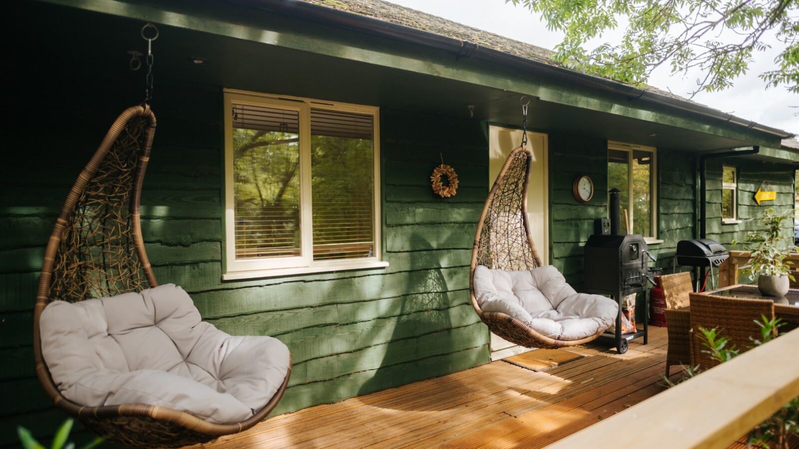 Two hanging chairs and a grill adorn the wooden deck of Chicken Shed Lodge, set against the charming backdrop of a green house with a window and door visible.