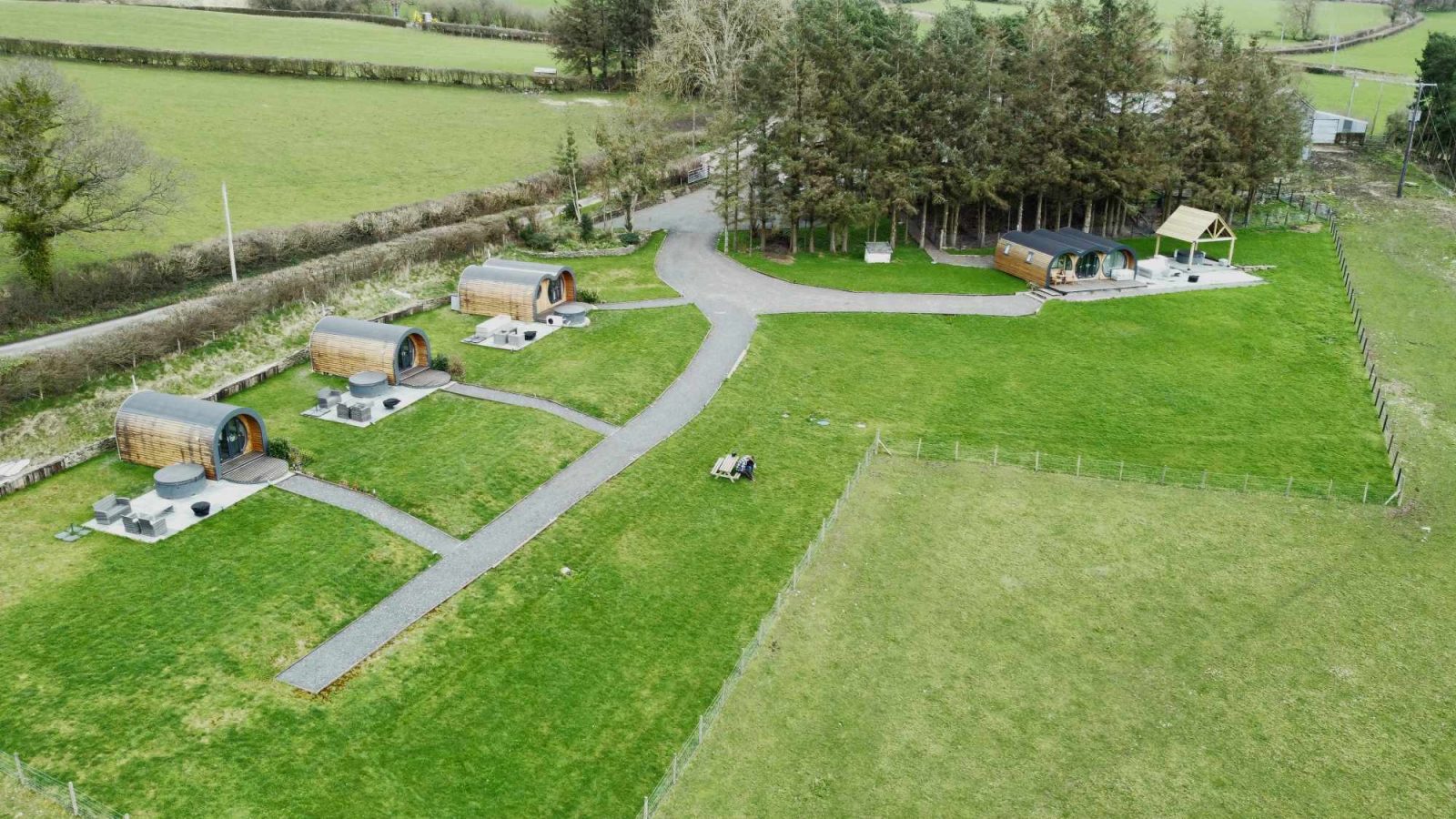 Aerial view of a grassy landscape featuring four wooden pod cabins in a semi-circle at Great House Farm. A larger cabin nestles near a cluster of trees, perfect for glamping. Paths connect the cabins, and fields and hedges surround the serene area.