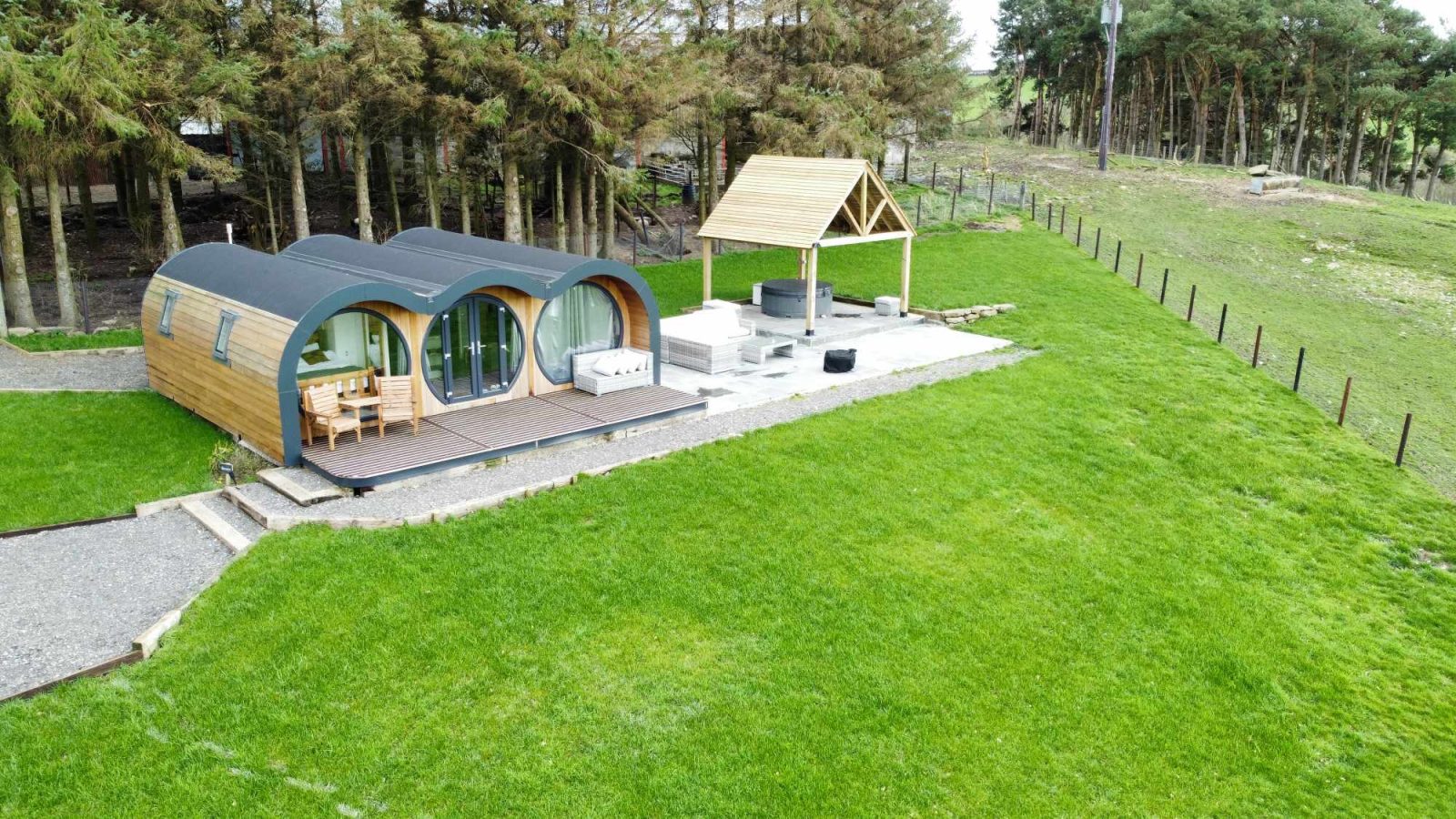 Aerial view of the triple cabin at Great House Farm Glamping with an arched roof, wooden deck, and outdoor seating, nestled in a grassy area near trees—an idyllic glamping experience.