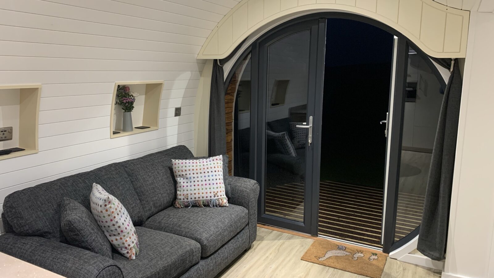 Modern living room with a dark gray sofa and patterned pillows, wooden floor, and large arched glass door leading to a deck, offering a touch of glamping luxury reminiscent of the serene escapes at Great House Farm.