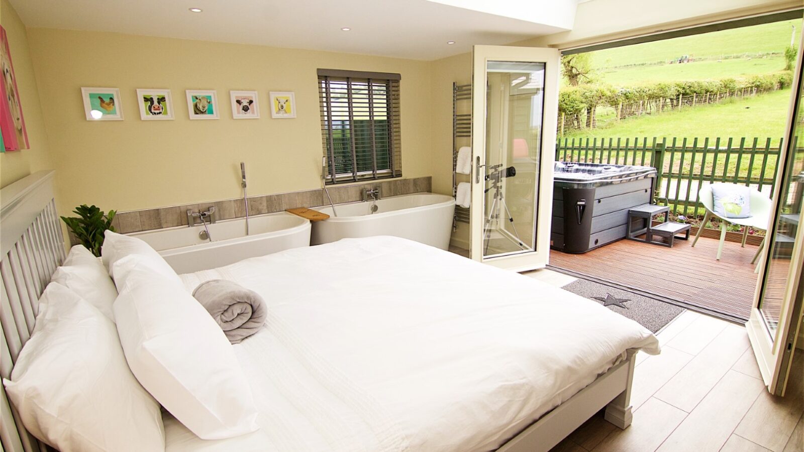 Bright bedroom with double bed, twin bathtubs, and doors opening to a patio with a hot tub under the shade of a tree and view of a grassy field.