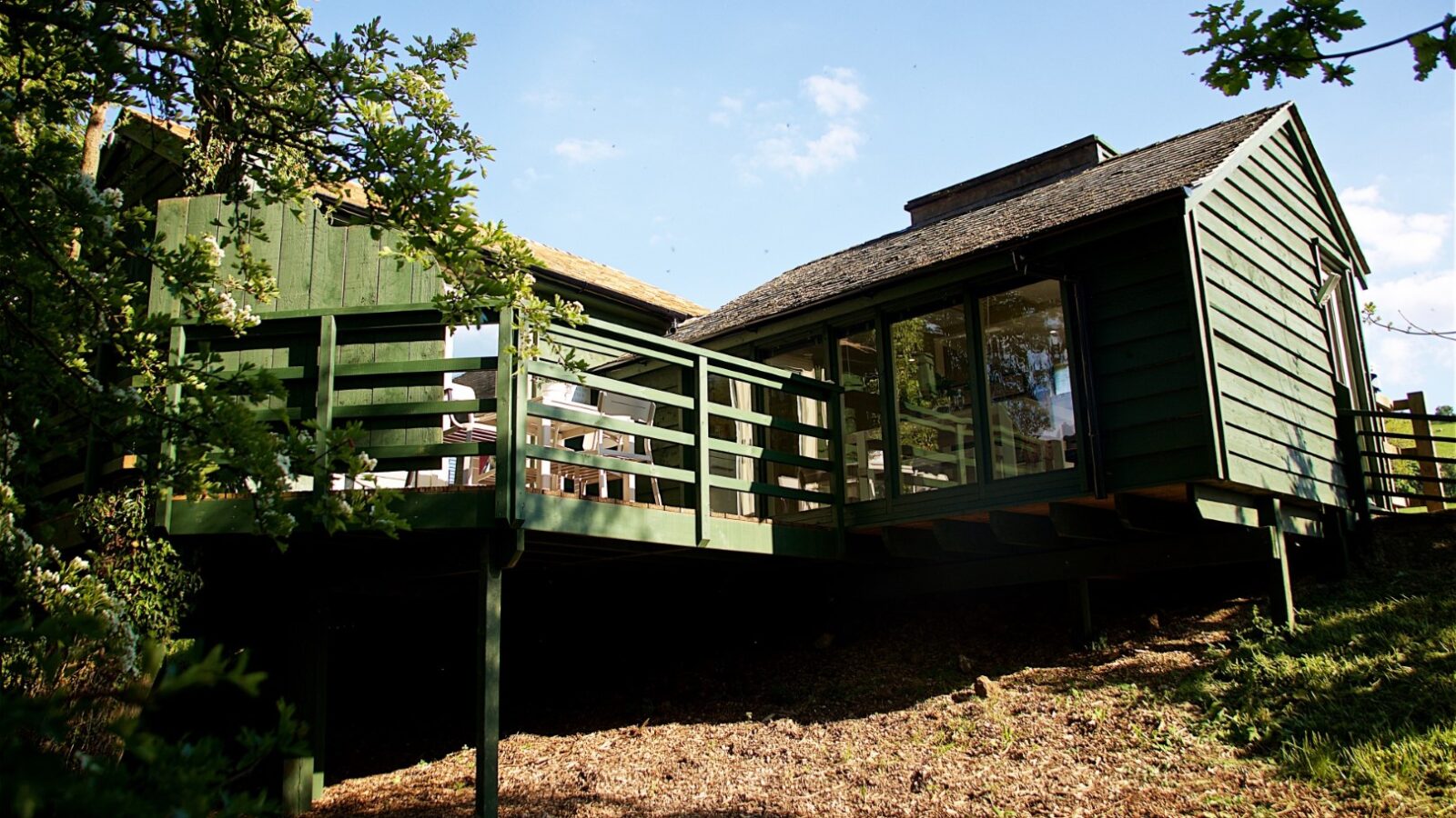 Jess’ Tree Bach: A modern green cabin with large windows and a wooden deck, this hillside treehouse retreat sits under a clear blue sky, offering the perfect vacation rental.