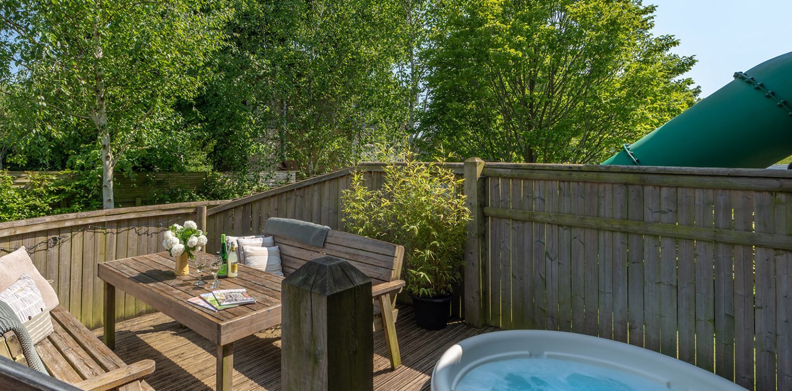 On Lavender Hill, a wooden deck features a table, bench, and sofa set beside a hot tub. Surrounded by lush greenery and treehouses, the area is enclosed by a wooden fence with a large green outdoor slide partially visible in the background.