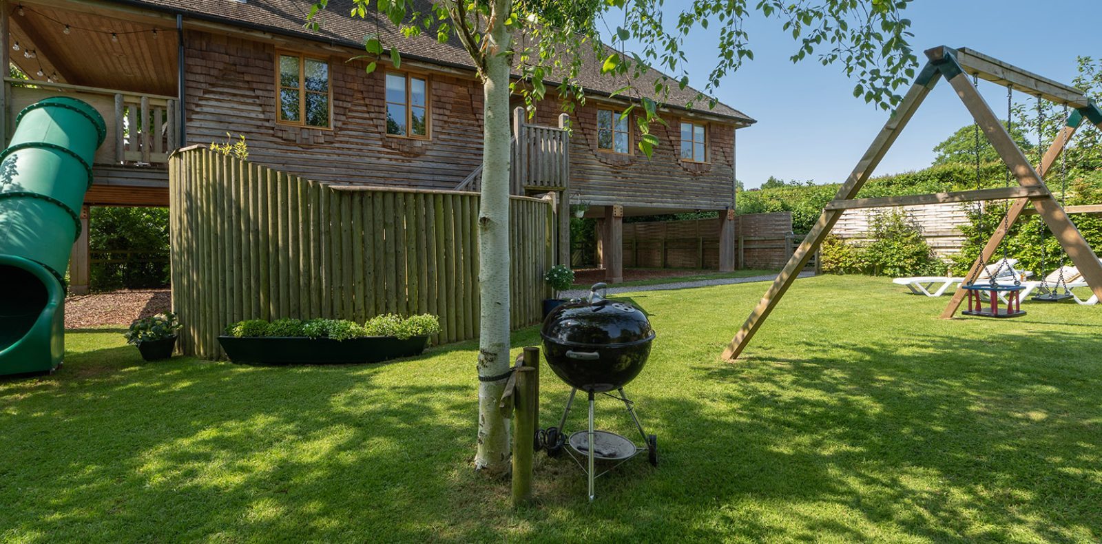 A lush backyard on Lavender Hill features a wooden play structure with a green slide and swing set. A charcoal grill stands near the treehouse, and a picnic table is visible in the background. The setting is sunny, with a wooden house partially visible.