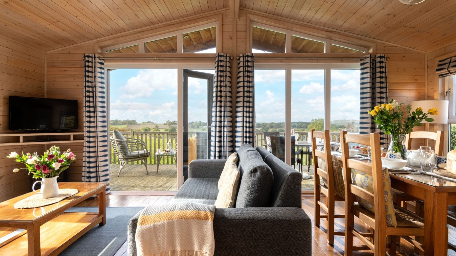 The cozy interior of this wooden cabin at Moonrise Lodges features a welcoming sofa, a charming dining table adorned with flowers, and offers stunning countryside views from the sunlit deck, ensuring unforgettable accommodations.