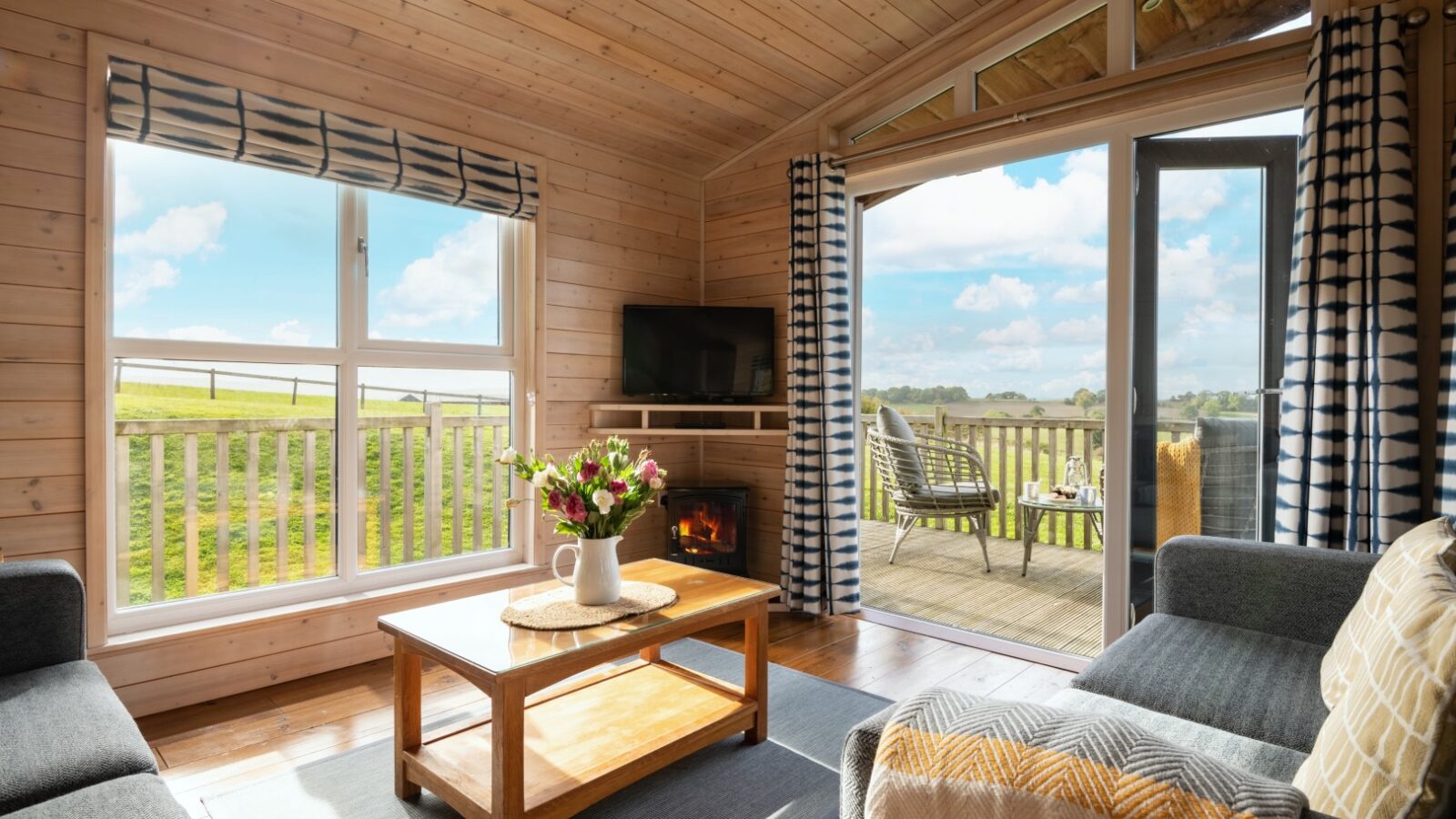 A cozy wooden living room with a fireplace, large windows, and a view of a deck overlooking green fields, reminiscent of charming lodges.