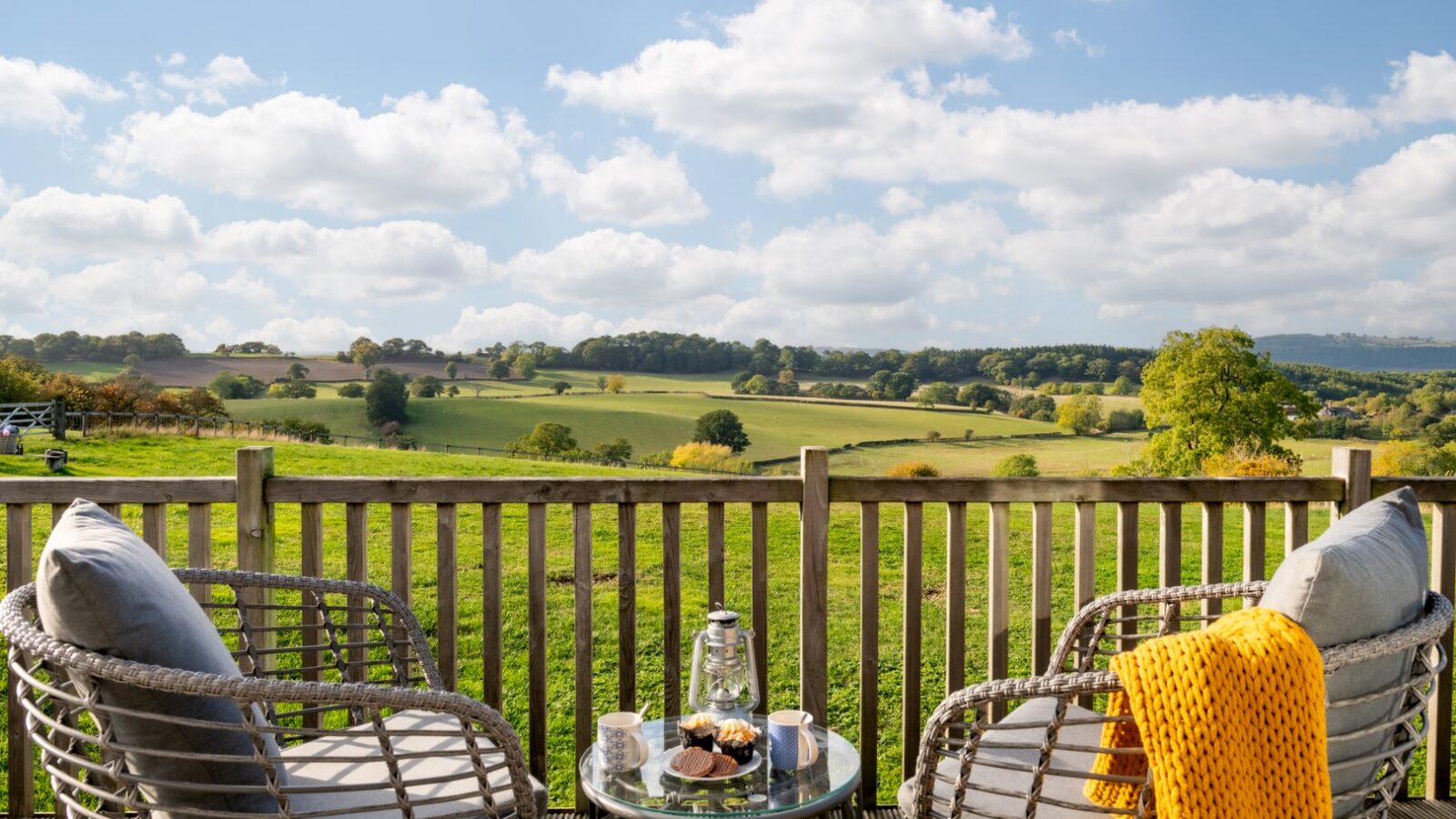 Two wicker chairs and a table with refreshments on a wooden deck overlook a scenic rural landscape under a partly cloudy sky, capturing the tranquil essence of Moonrise Lodges.