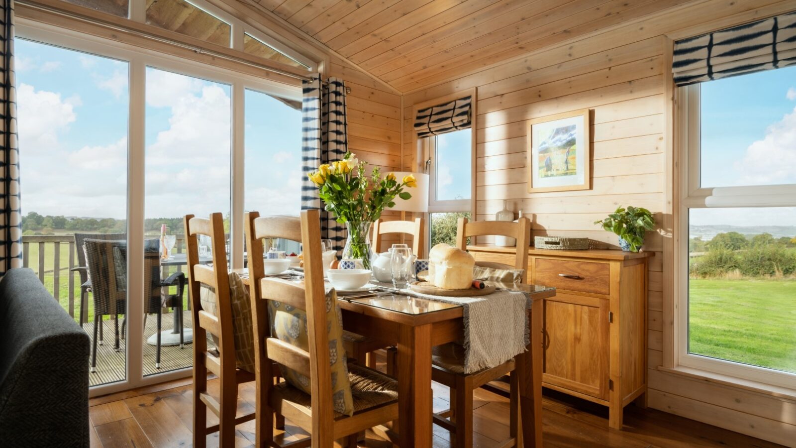 A bright wooden dining room at Moonrise Lodges features a table and chairs, perfectly positioned to offer a stunning view of the countryside through large windows.