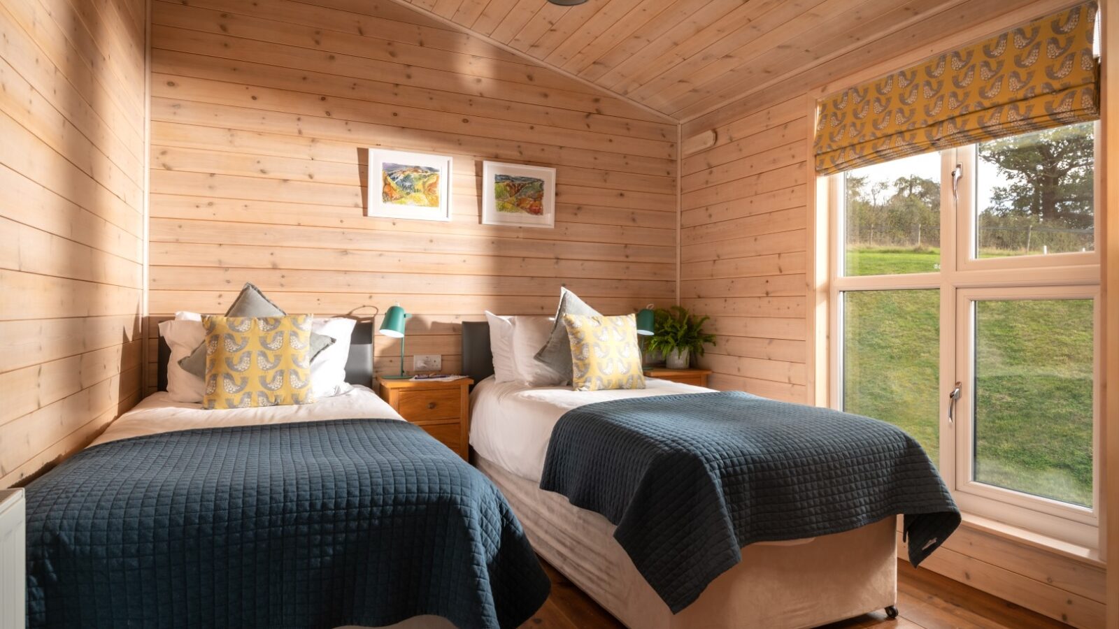 A cozy bedroom at Moonrise Lodges offers two single beds with blue blankets and patterned cushions. The wooden walls and a window reveal a scenic view, perfect for relaxation.