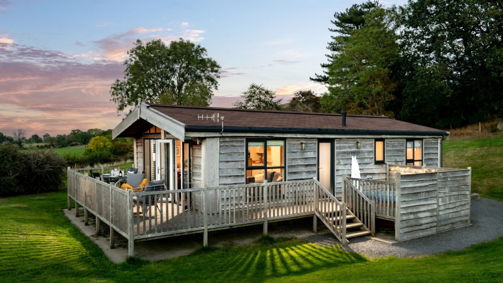 Moonrise Lodges features a charming wooden cabin with a wraparound deck, nestled on a grassy lawn and surrounded by trees, all set under a pink and blue evening sky.