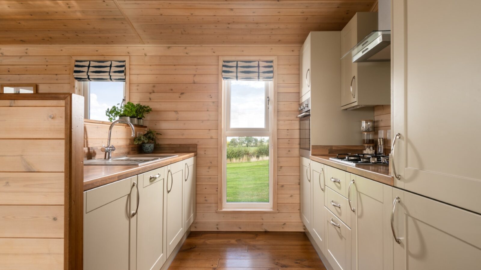 The wood-paneled kitchen, reminiscent of cozy lodges, features white cabinets and a window view of the grassy landscape. Modern appliances and a sleek sink rest on the wooden countertop, completing this inviting space where functionality meets charm.