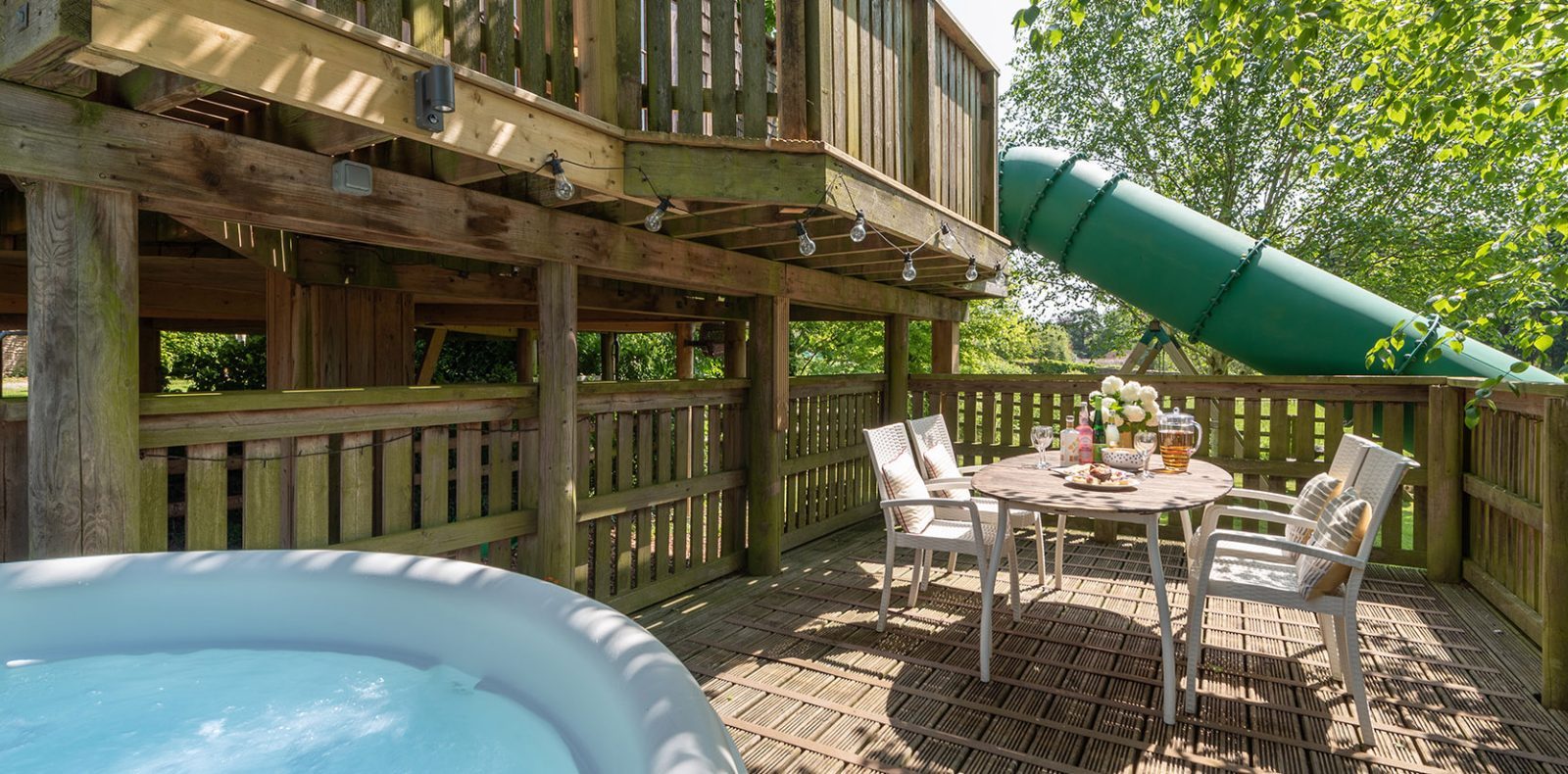 A cozy wooden deck with a round table and four chairs elegantly set with a vase of lavender sits under the serene ambiance of treehouses on the hill. Nearby, a hot tub beckons from its shaded spot, and a green tube slide descends from the upper level, surrounded by lush greenery.