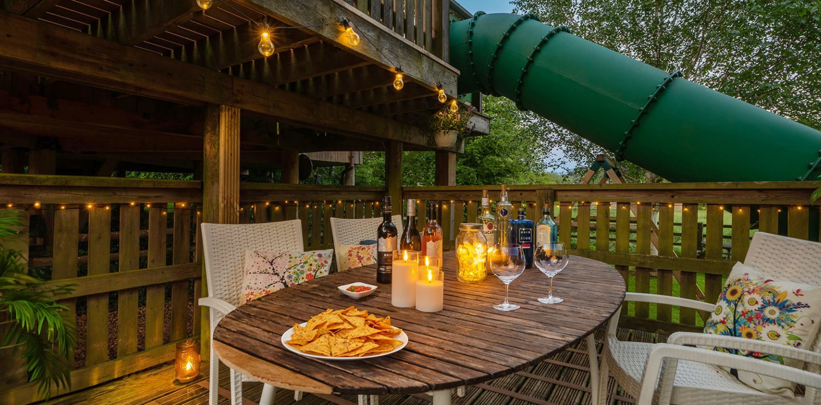 A cozy outdoor patio on Lavender Hill features a wooden table set with wine glasses, chips, and candlelight. White chairs surround it, and a green slide descends from the treehouse deck above. String lights add a warm glow to this enchanting evening setting.