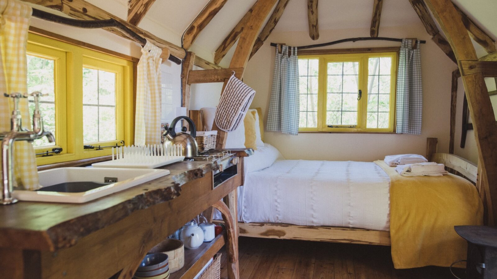 Cozy wooden cabin interior reminiscent of a woodcutter's retreat, featuring a comfortable bed, a simple kitchenette, and windows framed by charming yellow and plaid curtains.