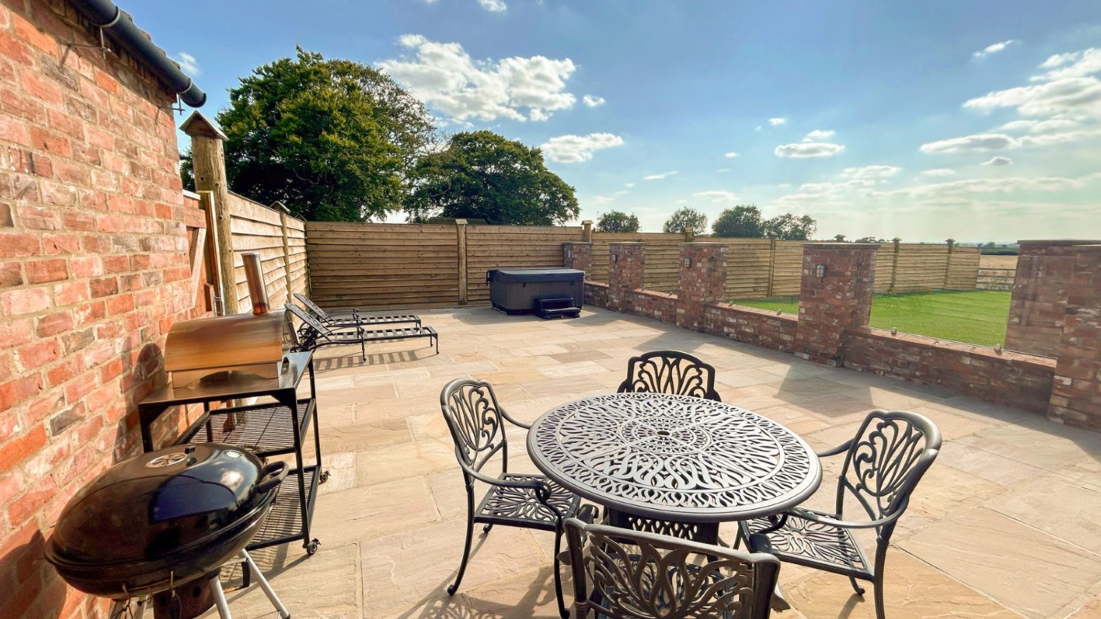 The Stables offers a sunny patio with a metal table and chairs, barbecue grill, sun loungers, and hot tub. Enjoy the charming ambiance with a brick wall and wooden fence providing a cozy backdrop.