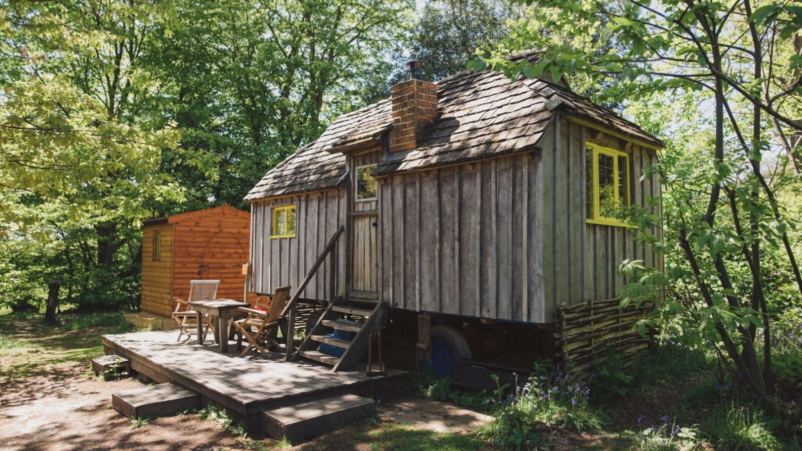 A rustic wooden woodcutters' cabin with a porch and outdoor seating, surrounded by trees and greenery, under a sunny sky.