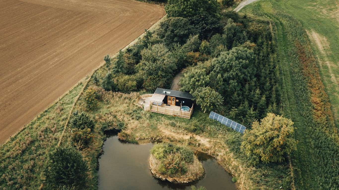 Aerial view of Duck Pond Cabin, featuring a cozy deck beside a tranquil pond. Surrounded by lush trees and sprawling fields, the cabin is complemented by nearby solar panels, harmonizing nature with sustainability.