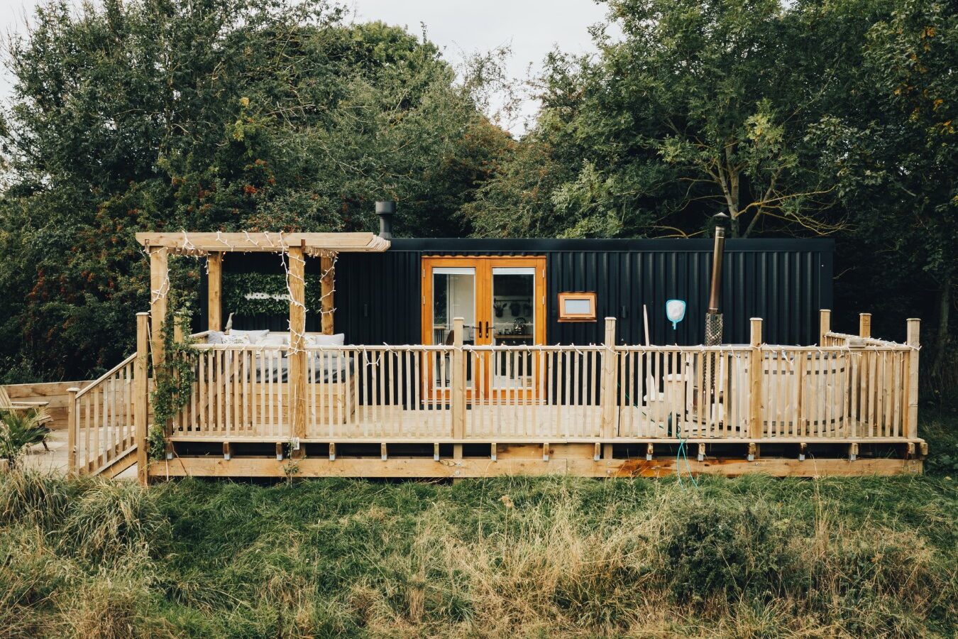 A small black cabin with a wooden deck and pergola, nestled near a peaceful duck pond, is surrounded by trees and grass.