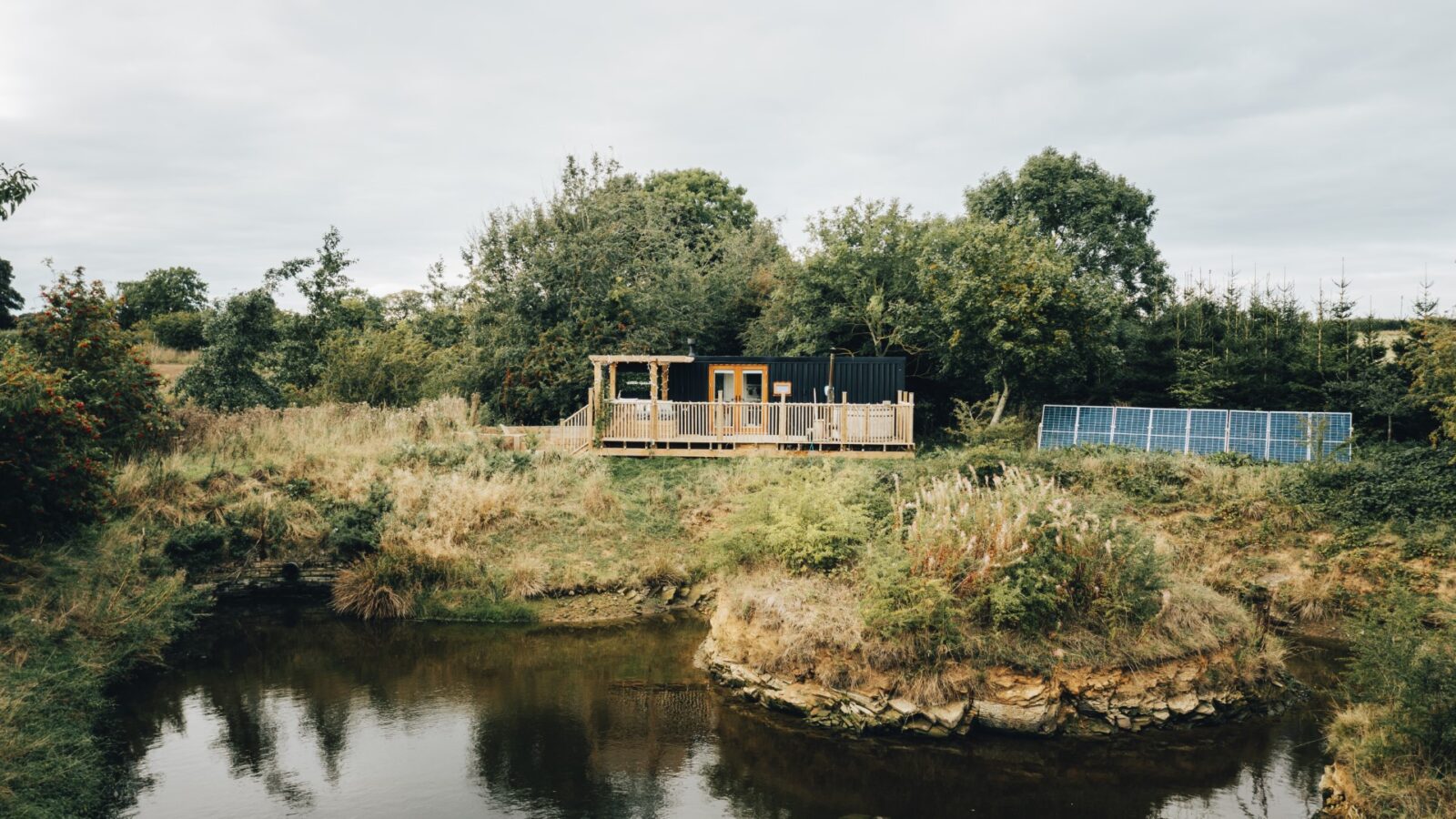 Duck Pond Cabin is a small house with a wooden deck that overlooks a pond, surrounded by trees and bushes. Solar panels are visible nearby.