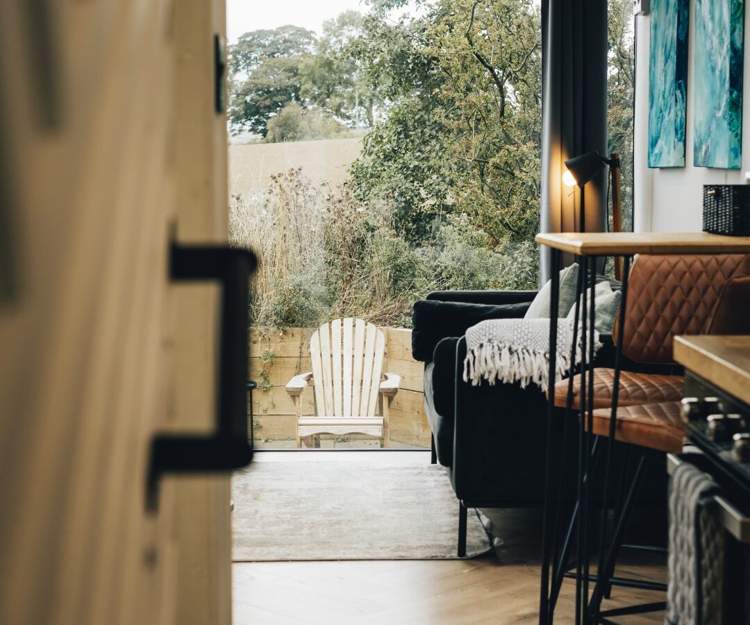 View from a cozy cabin room with plants outside, featuring a wooden chair on the patio, a sofa, bar stools, and a countertop. Just beyond the charming setup lies a serene duck pond that completes this tranquil retreat.