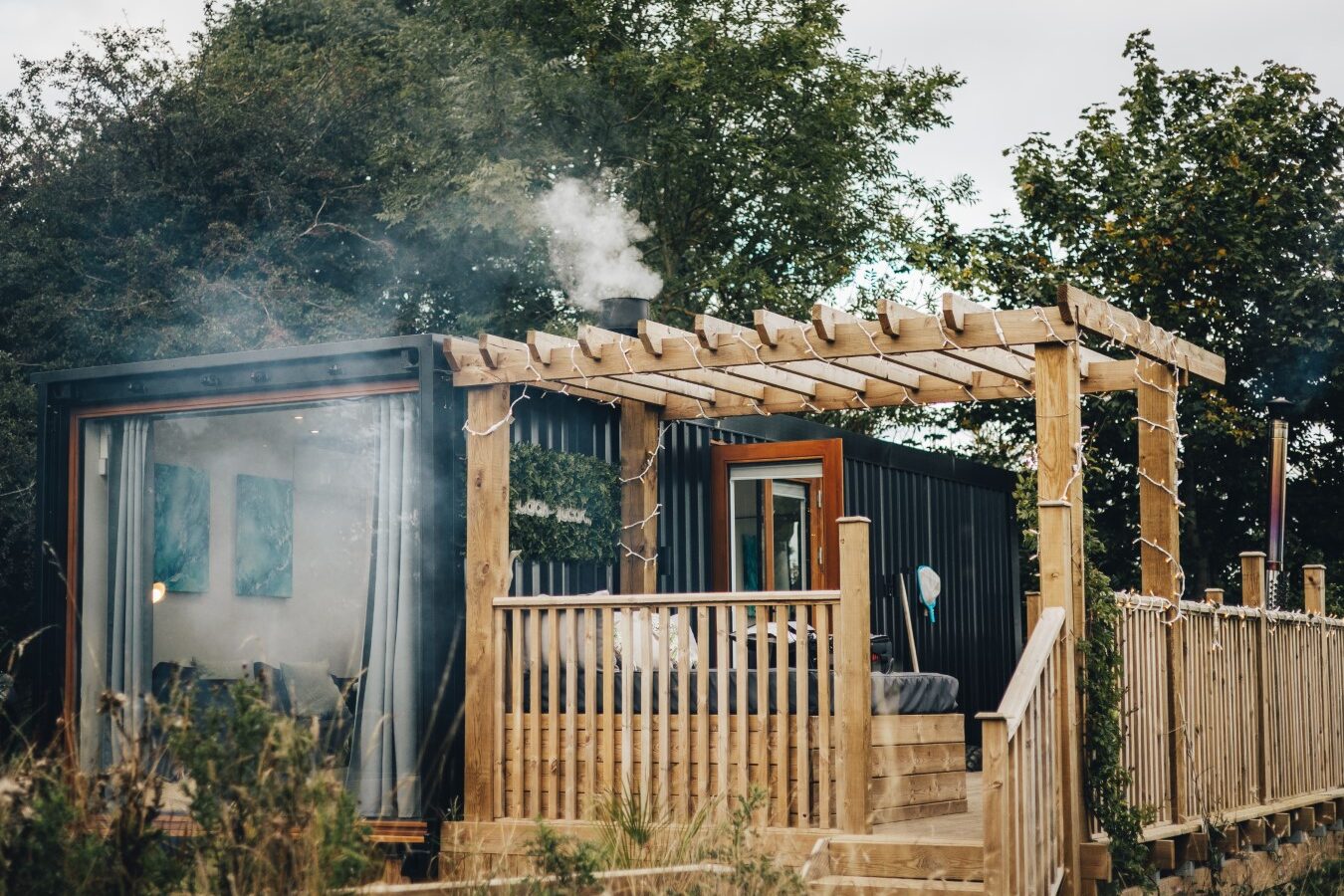 The Duck Pond Cabin is a modern tiny house with wood accents and large windows, nestled among trees. Smoke curls gently from the chimney on the roof, adding to its rustic charm.