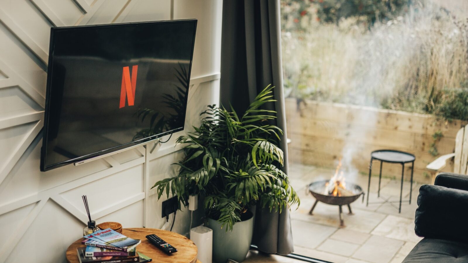 This cozy Duck Pond Cabin living room features a wall-mounted TV streaming Netflix, a table adorned with books and a remote, and an inviting outdoor area complete with a fire pit.