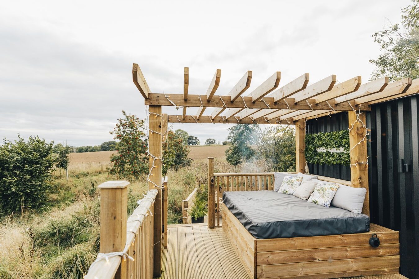 A charming wooden deck with a pergola and cushioned seating offers a serene view, overlooking a field with trees and a cloudy sky, evoking the peaceful vibe of a cabin retreat near a tranquil duck pond.