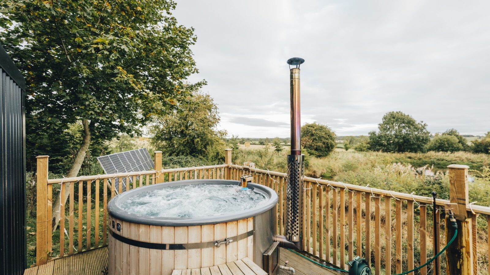 The wooden hot tub on a cozy deck offers a serene view of lush greenery and trees at Duck Pond Cabin, all under a moody, cloudy sky.