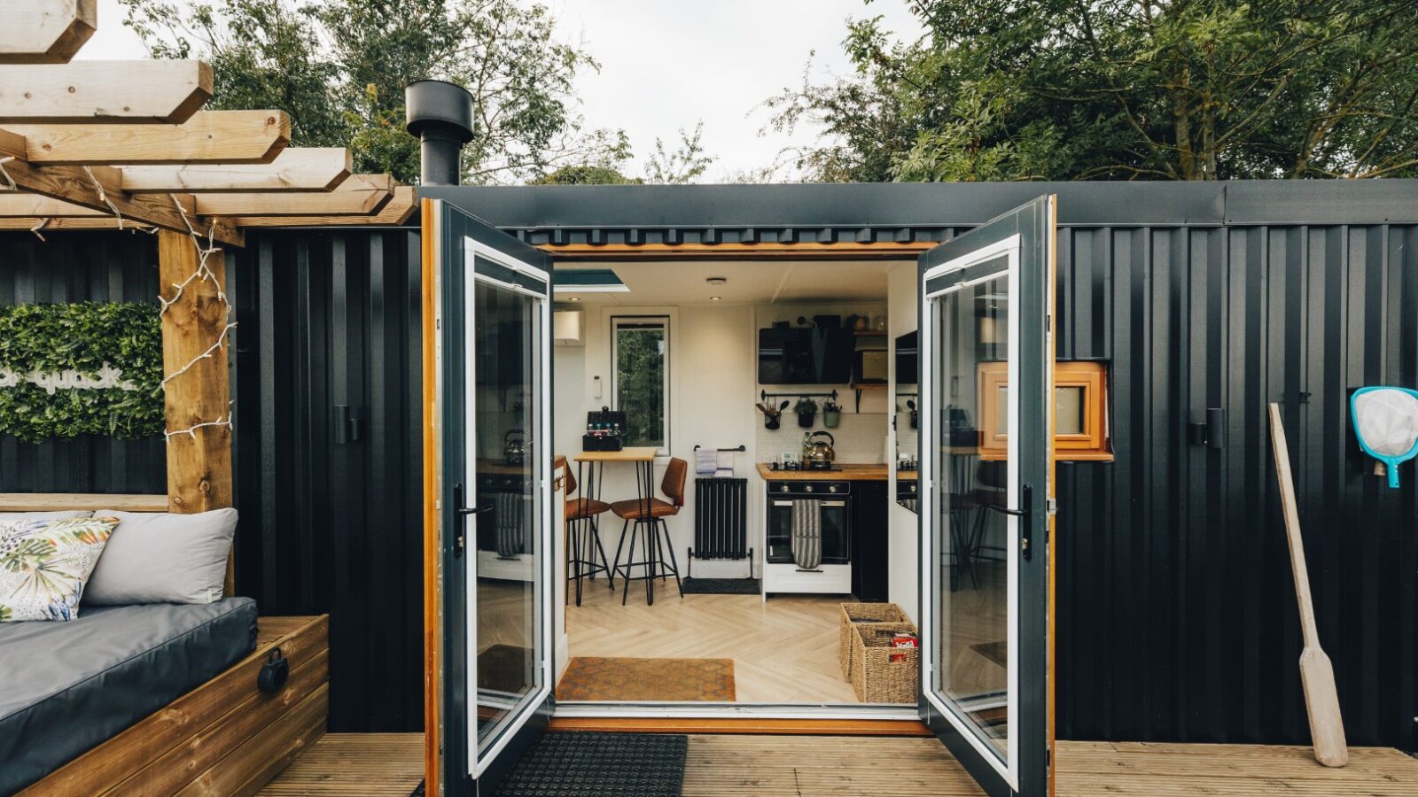 Open glass doors reveal the interior of a modern container home, known as Duck Pond Cabin, with a kitchen and stools, surrounded by trees and outdoor seating.