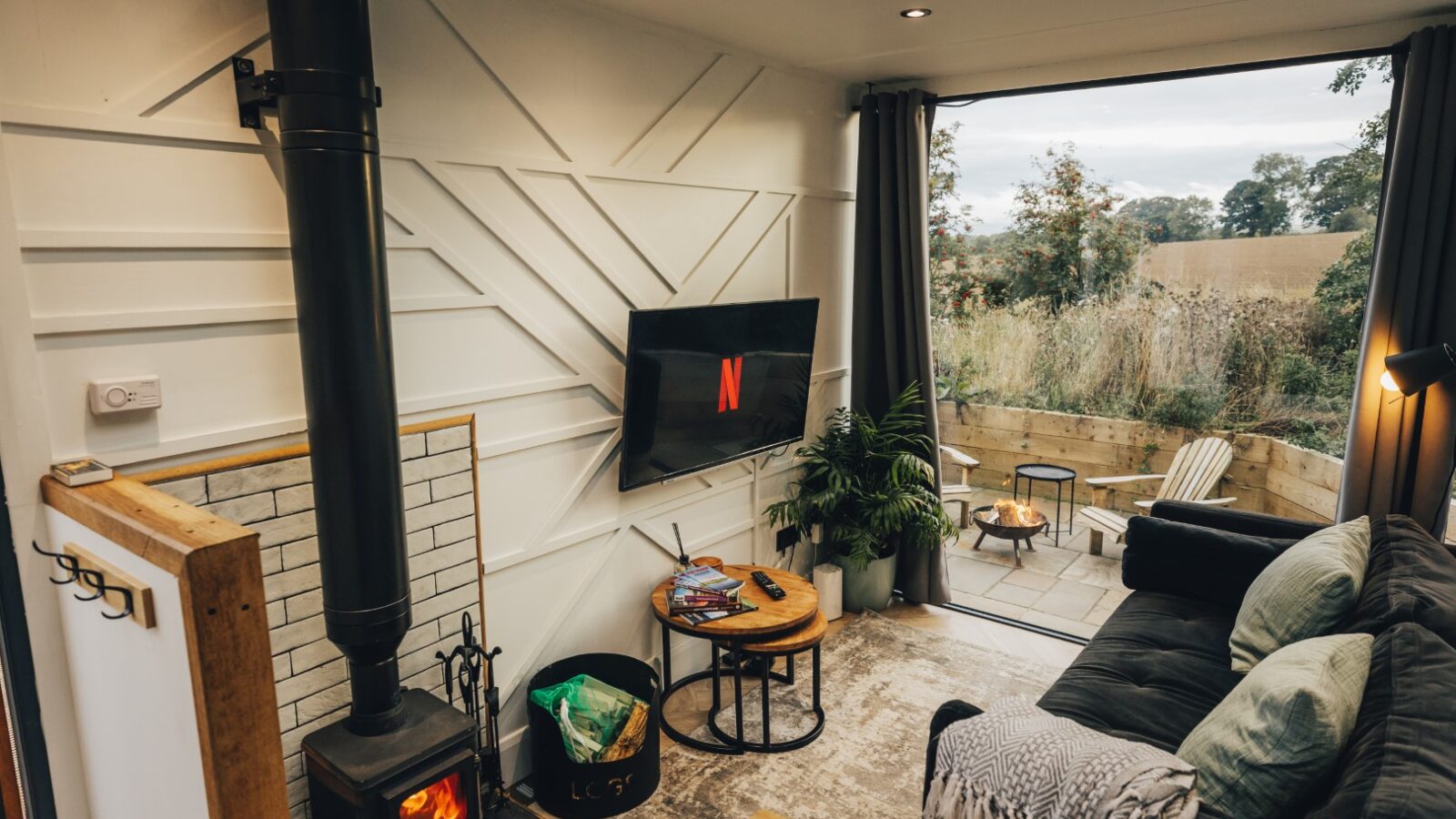 Cozy cabin living room with a wood stove, wall-mounted TV, sofa, plants, and a large window overlooking a deck with seating and views of the trees near the duck pond.