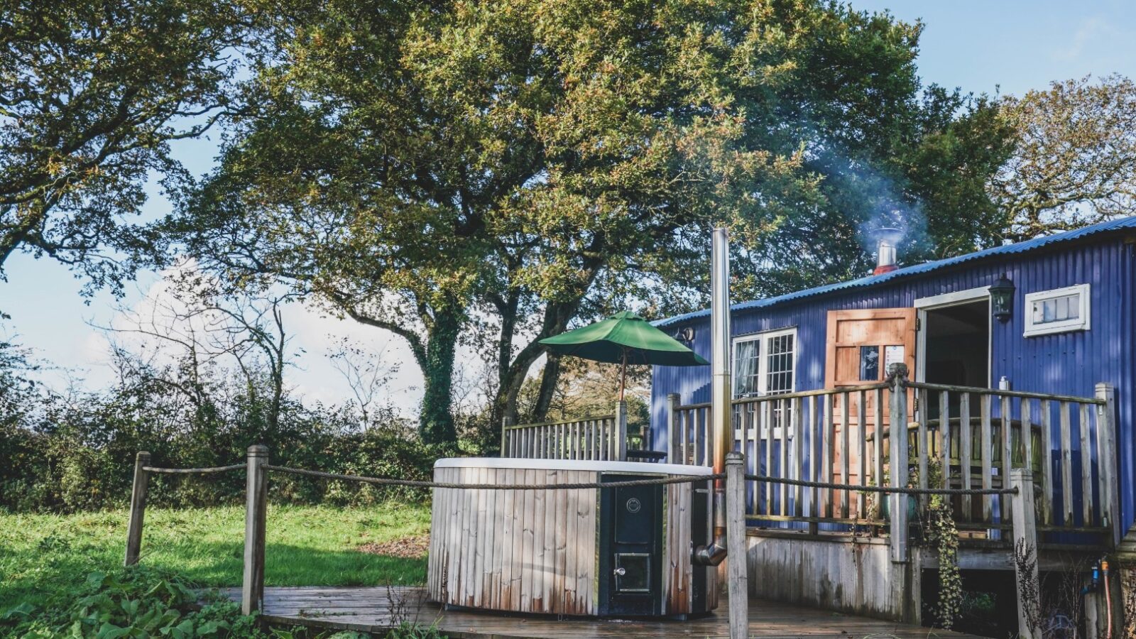 This wooden cabin at Leworthy Manor boasts blue walls, a deck, and a hot tub nestled in a grassy hideaway, surrounded by trees under a clear sky.